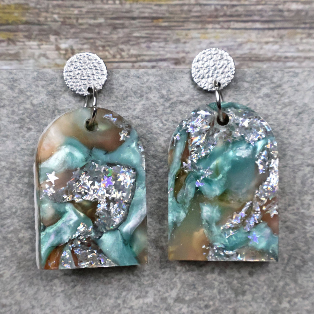 A pair of one of a kind arch shaped recycled acrylic earrings with silver, green and brown design titled 'Celestial Tide' and stainless steel toppers. Pair 102-02