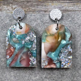 A pair of one of a kind arch shaped recycled acrylic earrings with silver, green and brown design titled 'Celestial Tide' and stainless steel toppers. Pair 102-05