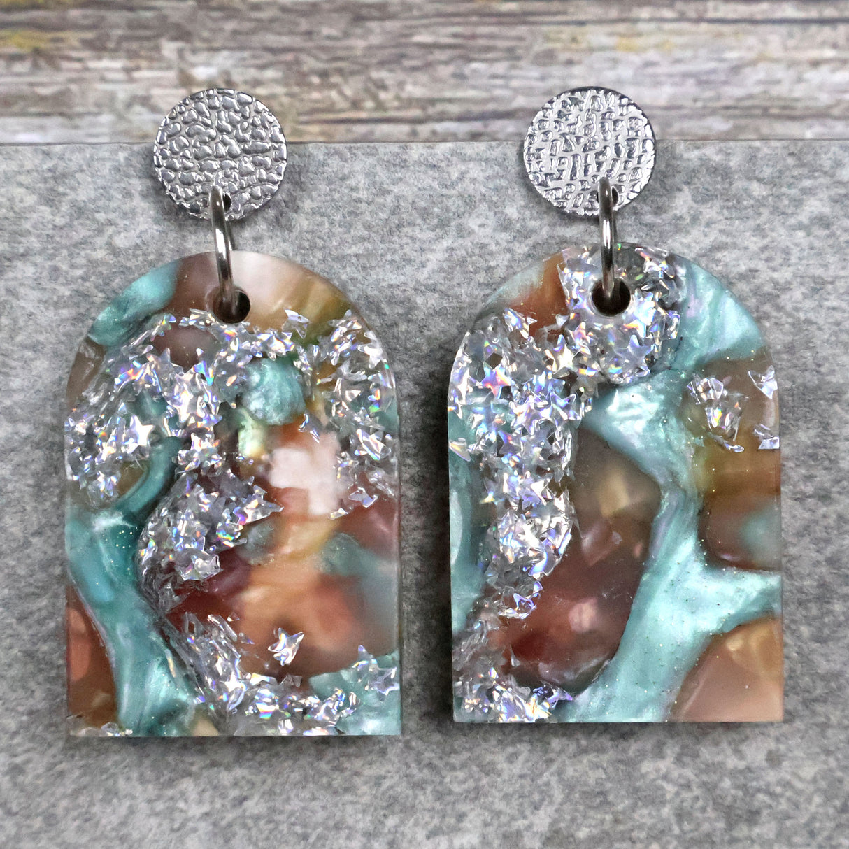 A pair of one of a kind arch shaped recycled acrylic earrings with silver, green and brown design titled 'Celestial Tide' and stainless steel toppers. Pair 102-08