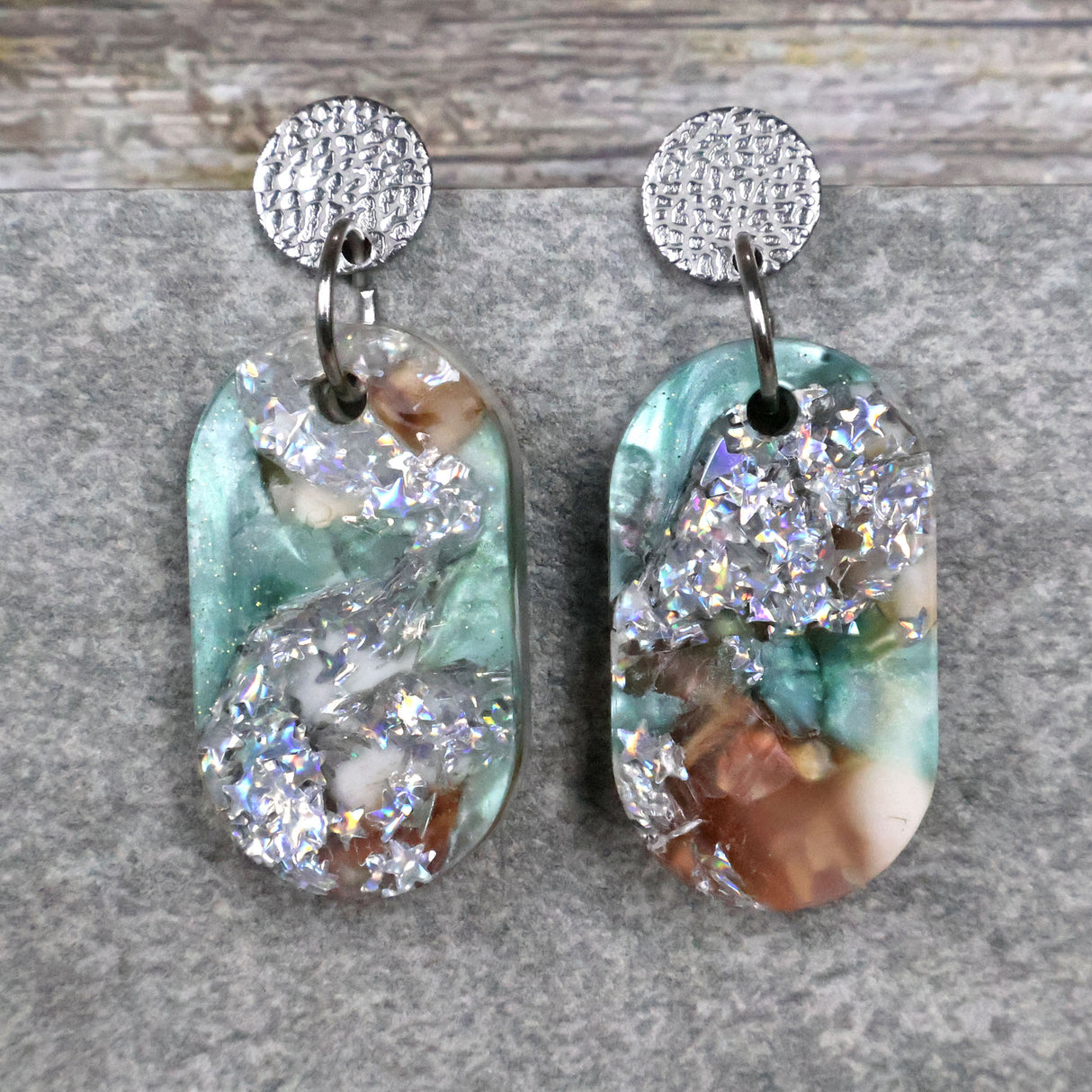 A pair of one of a kind oval shaped recycled acrylic earrings with silver, green and brown design titled 'Celestial Tide' and stainless steel toppers. Pair 102-09