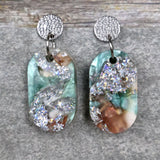 A pair of one of a kind oval shaped recycled acrylic earrings with silver, green and brown design titled 'Celestial Tide' and stainless steel toppers. Pair 102-09