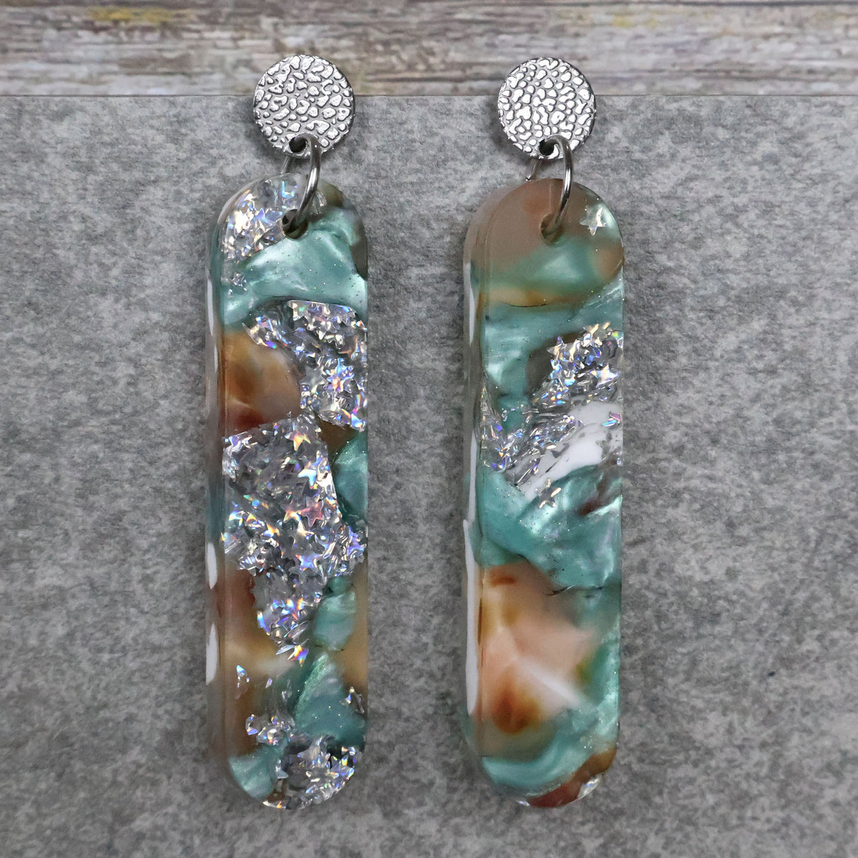A pair of one of a kind long oval shaped recycled acrylic earrings with silver, green and pink design titled 'Celestial Tide' and stainless steel toppers. Pair 102-12