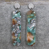A pair of one of a kind long oval shaped recycled acrylic earrings with silver, green and pink design titled 'Celestial Tide' and stainless steel toppers. Pair 102-12