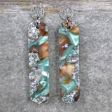 A pair of one of a kind long oval shaped recycled acrylic earrings with silver, green and brown design titled 'Celestial Tide' and stainless steel toppers. Pair 102-13