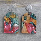 Pair of one of a kind arch shaped acrylic earrings with multicolored  design titled 'Sunset Reef' and stainless steel toppers on a gray background. Pair 106-01