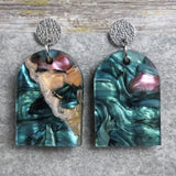 Pair of one of a kind arch shaped acrylic earrings with multicolored  design titled 'Sunset Reef' and stainless steel toppers on a gray background. Pair 106-02