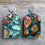 Pair of one of a kind arch shaped acrylic earrings with multicolored  design titled 'Sunset Reef' and stainless steel toppers on a gray background. Pair 106-03