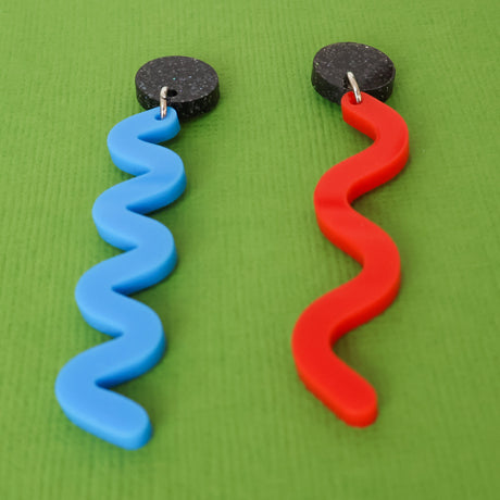 Pair of asymmetrical acrylic earrings with blue and red wavelength designs on a green background, angled view