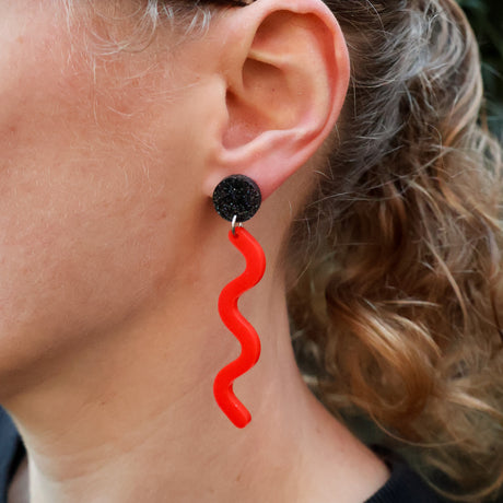 Closeup of a person wearing acrylic red wavelength earrings, hanging from a black glittery acrylic topper. 