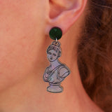 A close-up of a person's ear wearing a dangling earring. The earring has a dark black round stud and a silver-tone pendant shaped like a classical bust with black line art.
