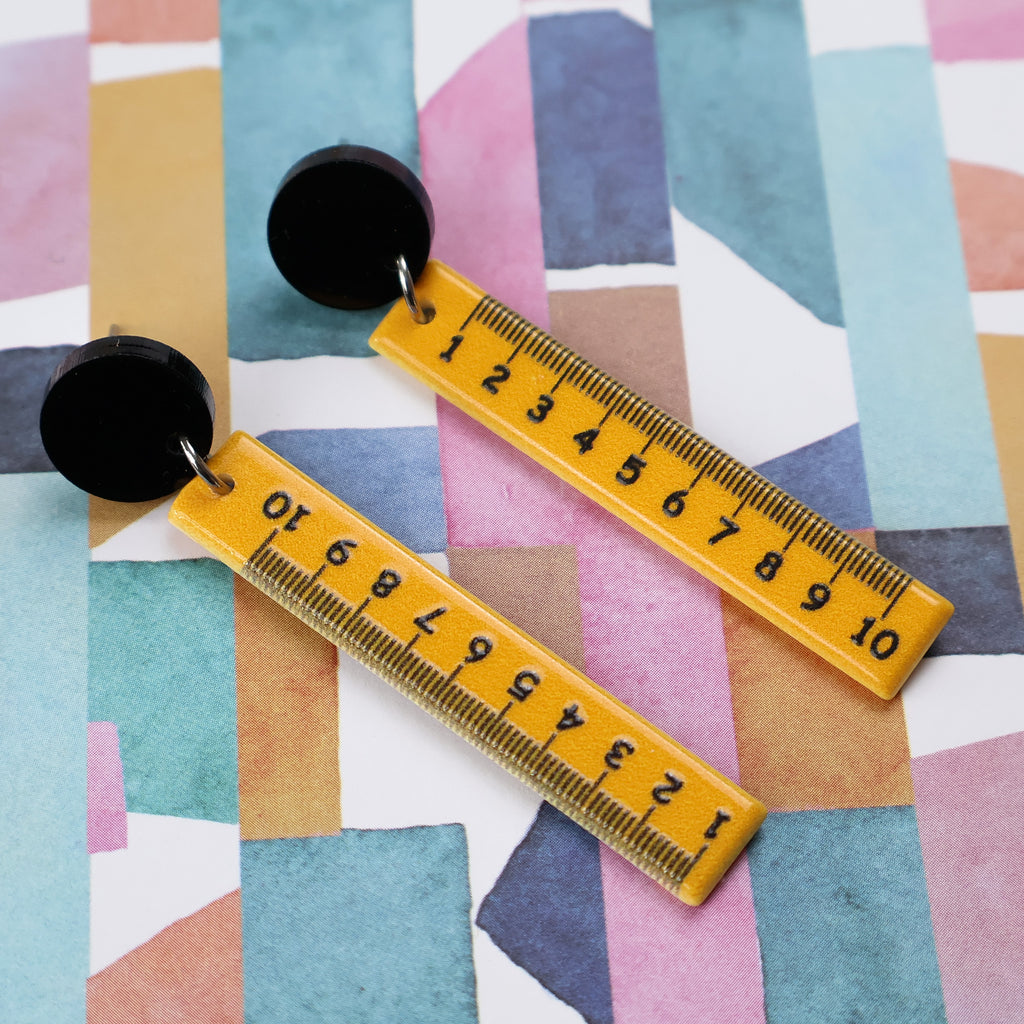 Yellow measuring ruler dangle earrings, hanging from black round acrylic toppers. 