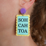 Earring with 'SOH CAH TOA' text on a light blue background, yellow border and purple topper, worn by a person.