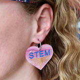 Swirly pink acrylic heart earrings with blue STEM text and a gold starburst pattern, hanging from stainless steel toppers. Earrings being modelled.