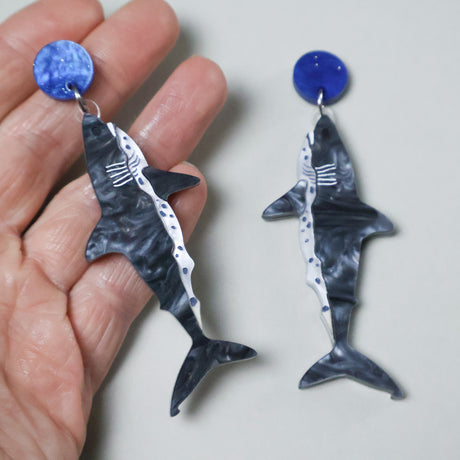 Shark-shaped acrylic earrings with blue tops held by a hand.