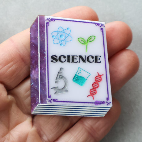 Acrylic brooch textbook with 'SCIENCE' and scientific symbols held in a hand against a neutral background