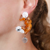 Acrylic dangle earring with a design of a sea otter and a shell hanging from it, worn by a person.