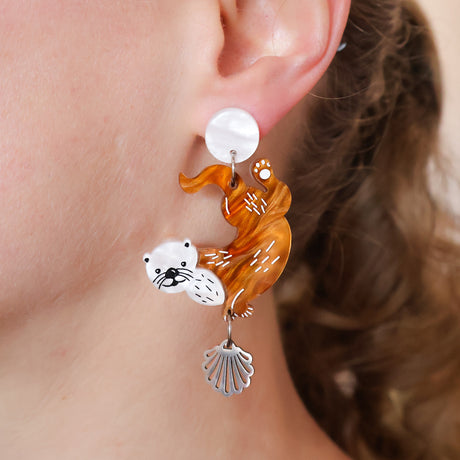Acrylic dangle earring with a design of a sea otter and a shell hanging from it, worn by a person.