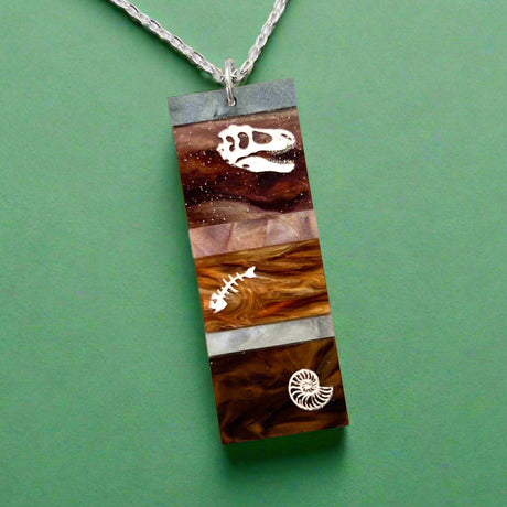 A pendant representing sedimentary layers, rectangular in shape and comprising 6 strips of different grey and brown acrylics, with a t-rex skull, fish bones and nautilus engraved and handpainted in the larger layers. The pendant hangs on a fine stainless steel chain.