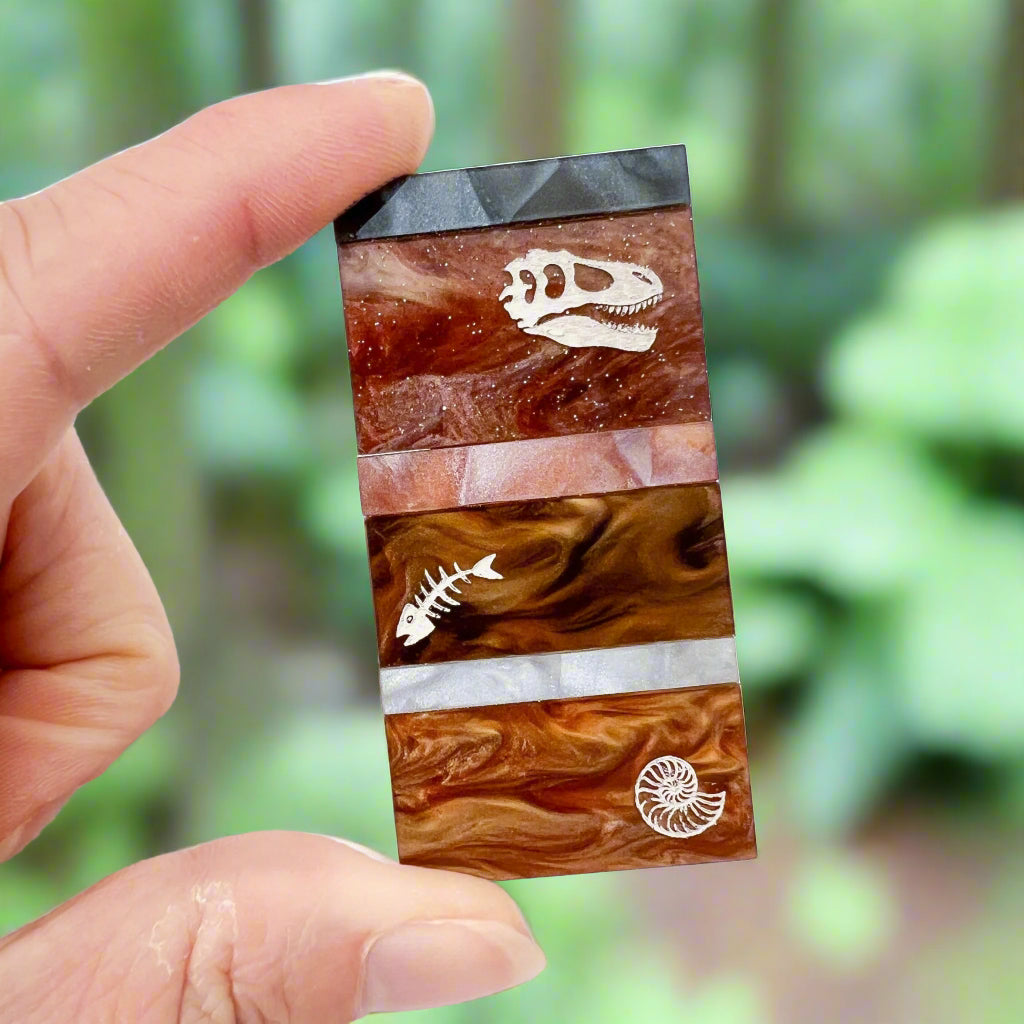 A brooch representing sedimentary layers, rectangular in shape and comprising 6 strips of different grey and brown acrylics, with a t-rex skull, fish bones and nautilus engraved and handpainted in the larger layers.