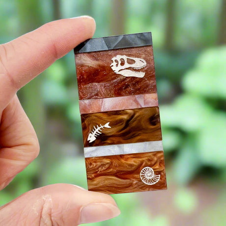 A brooch representing sedimentary layers, rectangular in shape and comprising 6 strips of different grey and brown acrylics, with a t-rex skull, fish bones and nautilus engraved and handpainted in the larger layers.