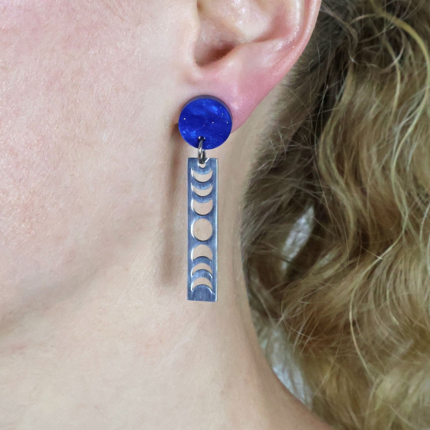 Earring with blue circular top and silver moon phase bar hanging from an ear