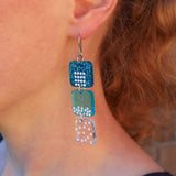 A close-up of a person wearing a dangling earring made in acrylic and representing the solid liquid and gas states of matter. Has french hooks.