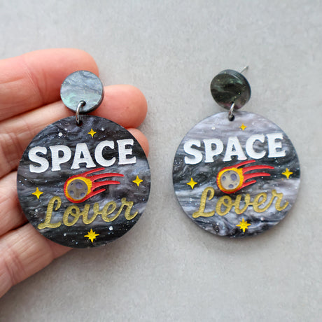 Earrings with 'Space Lover' design on a gray background with a meteor, being held in a hand