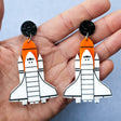 Laser cut acrylic space shuttle earrings in white, orange and black being held in a hand