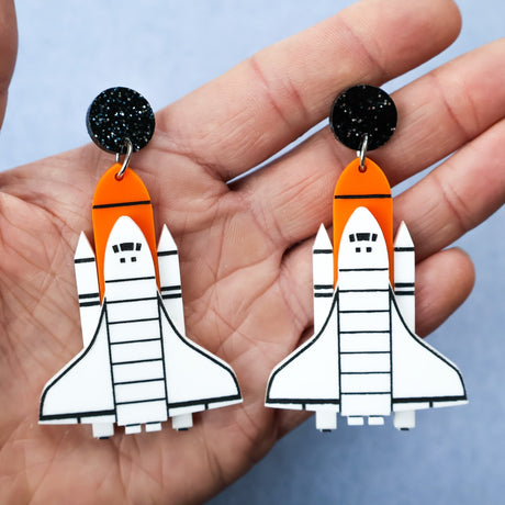 Laser cut acrylic space shuttle earrings in white, orange and black being held in a hand