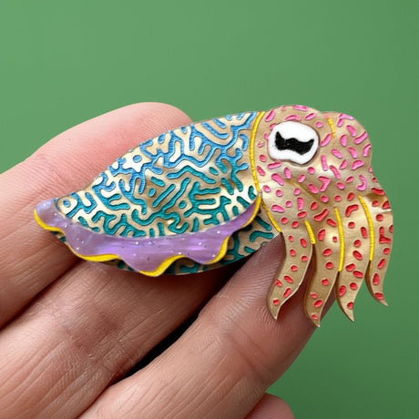 Colorful cuttlefish-shaped pin held between fingers against a green background