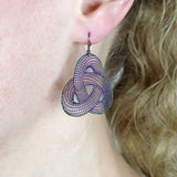 Close-up of a person wearing geometric surface themed earrings in a a rainbow toned stainless steel with french hook tops. 