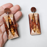 A pair of acrylic earrings displaying stalactite and stalagmites, made from brown and gold toned acrylics, and being held in a hand