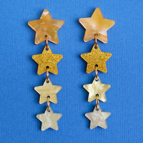 A pair of acrylic star stack earrings, featuring 4 different star shapes in different yellow and gold acrylics against a blue backdrop. 