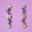 A pair of star and moon acrylic dangle earrings shown front on against a pink cardstock backdrop. The earrings consist of alternating small yellow moon and grey/brown stars. 