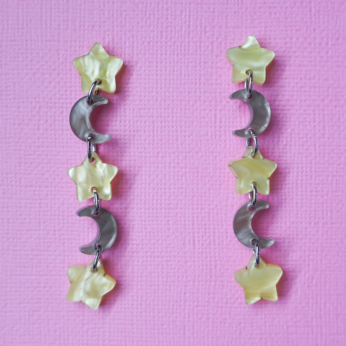A pair of star and moon acrylic dangle earrings shown front on against a pink cardstock backdrop. The earrings consist of alternating small yellow moon and grey/brown stars. 