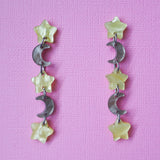 A pair of star and moon acrylic dangle earrings shown front on against a pink cardstock backdrop. The earrings consist of alternating small yellow moon and grey/brown stars. 