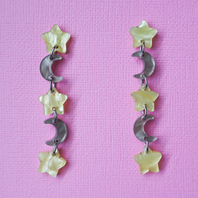 A pair of star and moon acrylic dangle earrings shown front on against a pink cardstock backdrop. The earrings consist of alternating small yellow moon and grey/brown stars. 