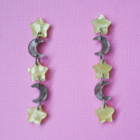 A pair of star and moon acrylic dangle earrings shown front on against a pink cardstock backdrop. The earrings consist of alternating small yellow moon and grey/brown stars. 