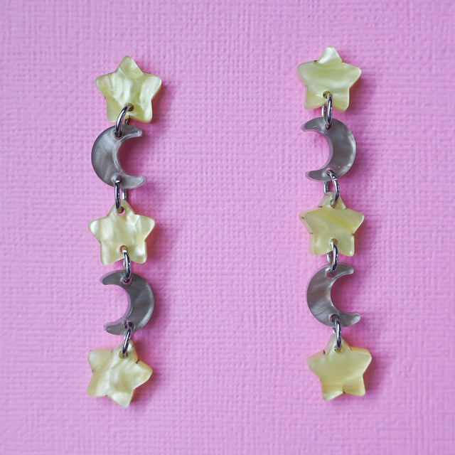 A pair of star and moon acrylic dangle earrings shown front on against a pink cardstock backdrop. The earrings consist of alternating small yellow moon and grey/brown stars. 