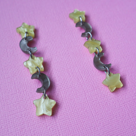 A pair of star and moon acrylic dangle earrings photographed at an angle. The earrings consist of alternating small yellow moon and grey/brown stars. 
