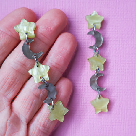 A pair of star and moon acrylic dangle earrings being held up in a hand. The earrings consist of alternating small yellow moon and grey/brown stars. 
