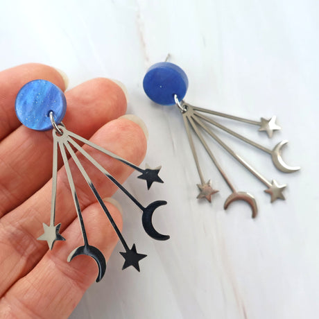 Earrings with blue round toppers and star and moon stainless steel dangles being held in a hand against a white background.