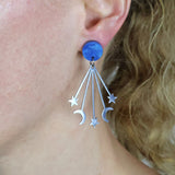 Stainless steel dangles with a moon and stars design and a blue acrylic  round topper, being modelled on an ear. 