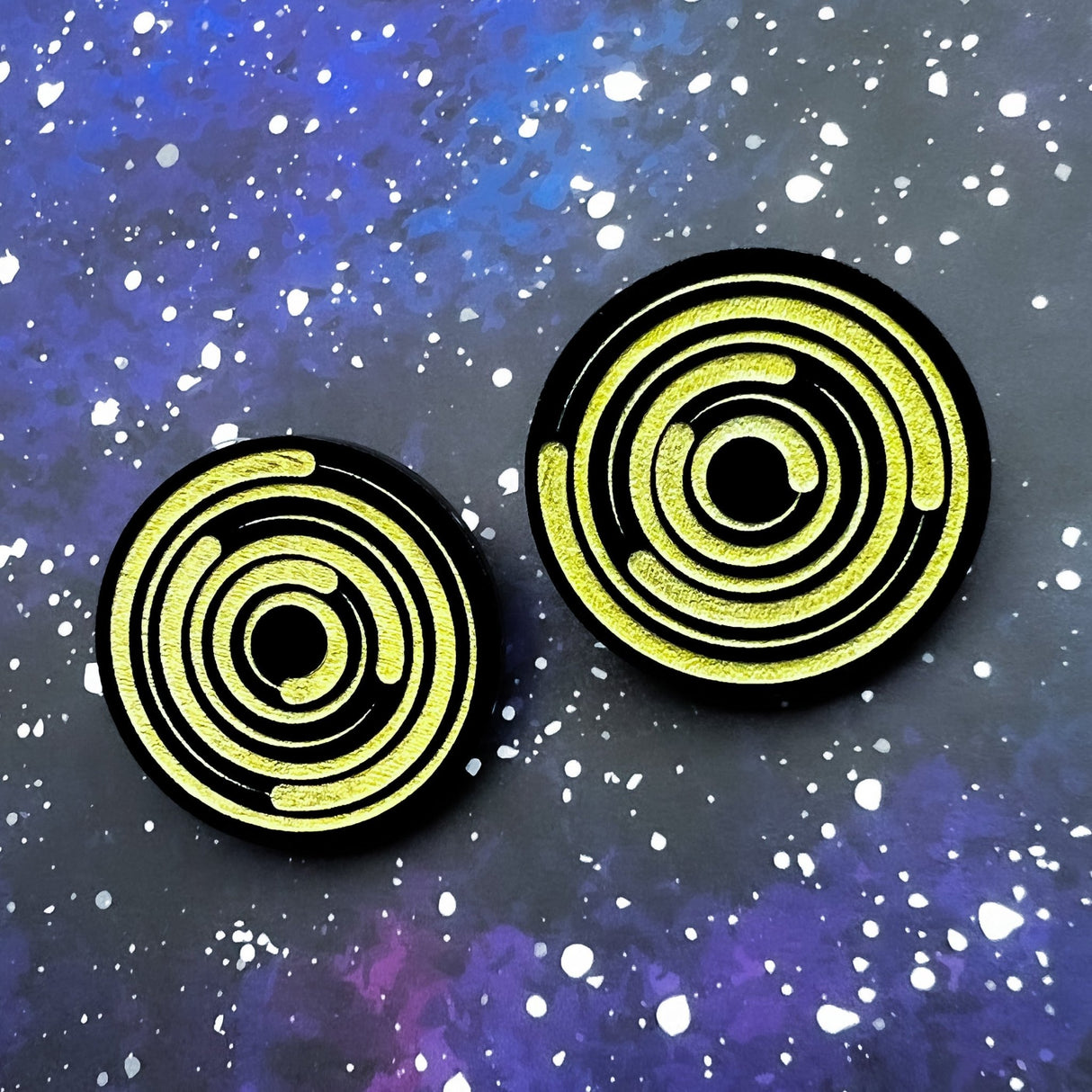 A pair of round acrylic star trail studs, black with gold painted star trails within. 