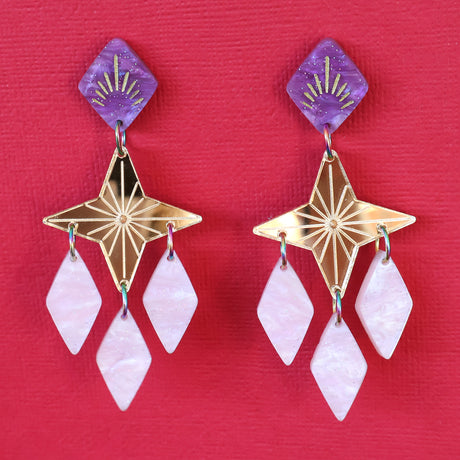 Gold pink and purple statement dangle earrings with a star focal piece against a deep red backdrop. Front View