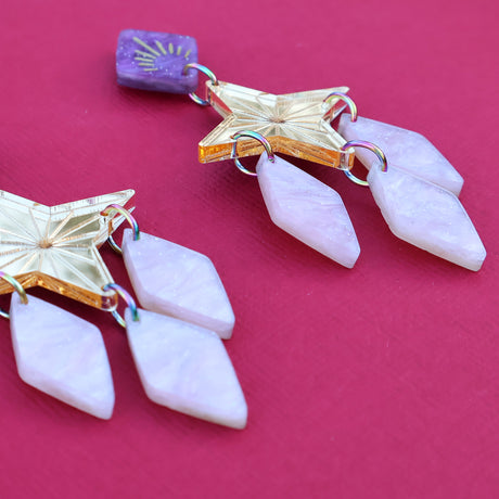 Gold pink and purple statement dangle earrings with a star focal piece against a deep red backdrop. Angled 