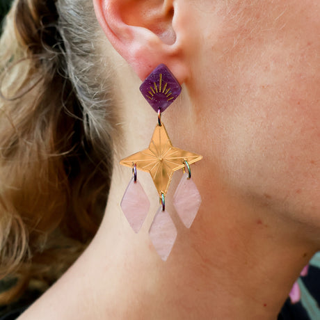 Close-up of a person wearing statement gold star dangle earring with purple and pink acrylic components. 