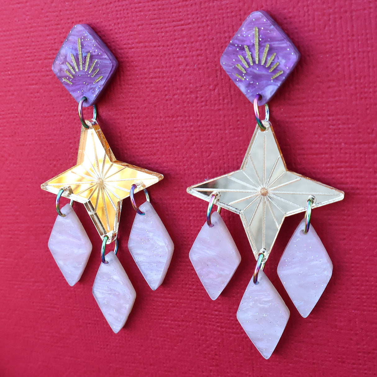 Gold pink and purple statement dangle earrings with a star focal piece against a deep red backdrop. Side angled view