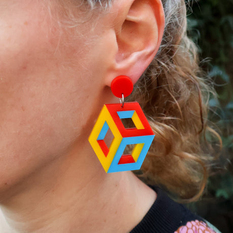 Close-up of a person wearing colorful geometric cube themed earrings in red blue and yellow tones.
