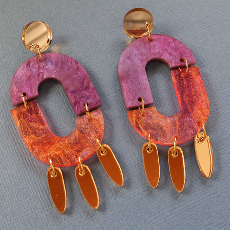 Pair of colourful dangle earrings in pinks and purples with gold accents on a gray background